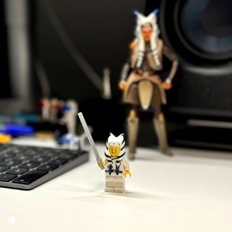 Close-up of a LEGO knight minifigure holding a sword on a desk, second blurred figure and speaker in the background, keyboard and office surface visible