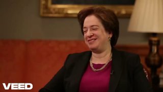 Woman in a dark jacket smiling while seated on a couch in an interview setup, warm lighting, lamp and framed art behind her.