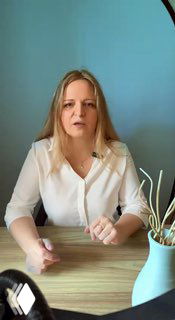 Женщина-лектор за столом на фоне голубой стены, объясняет бухгалтерские и налоговые нюансы в формате видеоролика для предпринимателей