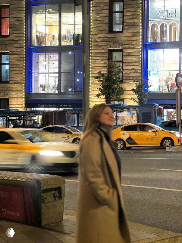 Женщина в бежевом пальто идёт по ночной городской улице мимо ярко освещённых витрин; на фоне видны проезжающие жёлтые такси и динамика улицы.