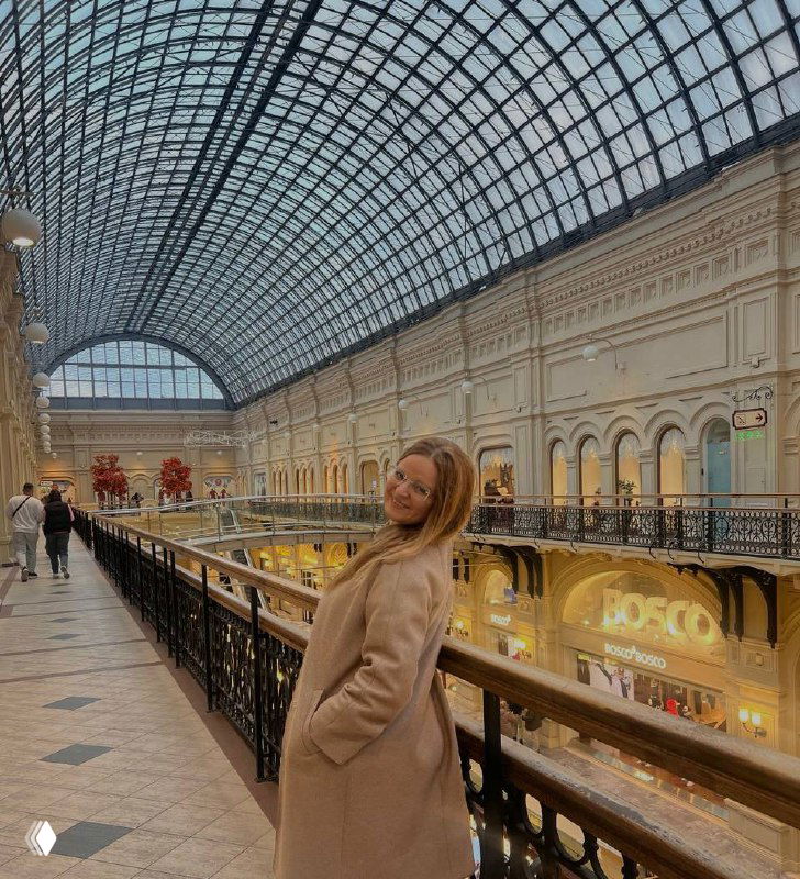 Женщина в бежевом пальто стоит на балконе большого торгового центра под высоким стеклянным куполом; вокруг витрины и галереи магазинов в тёплом освещении.