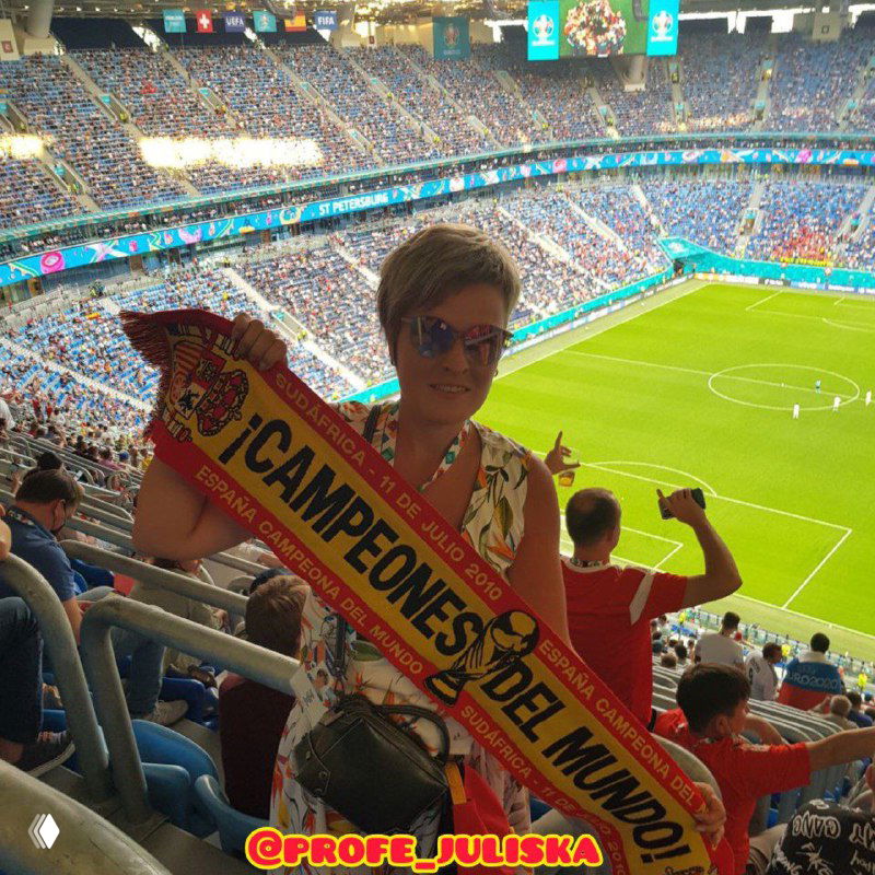 Женщина на трибуне переполненного стадиона держит шарфик сборной Испании с надписью «¡Campeones del Mundo!», виден футбольный газон и болельщики.