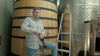 Мужчина в винном погребе держит бутылку кавы на фоне большой деревянной бочки, стальных резервуаров и лестницы