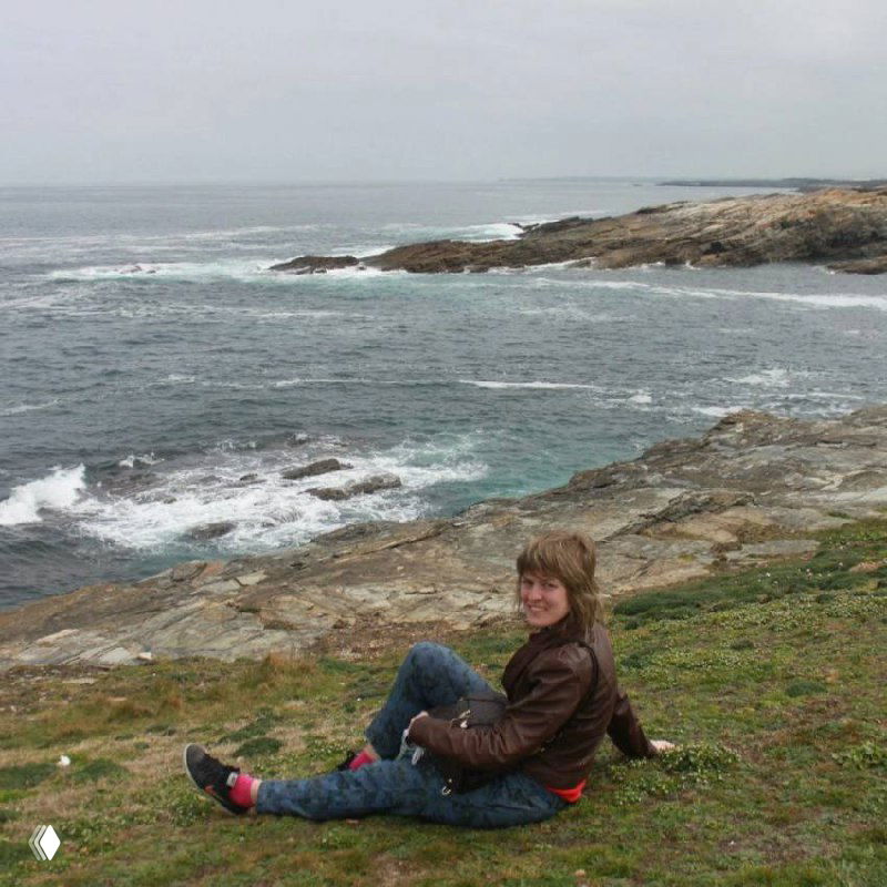 Автор сидит на скалистом берегу Атлантического океана в Галисии; видны волны, камни и пасмурное небо, атмосферный морской пейзаж.