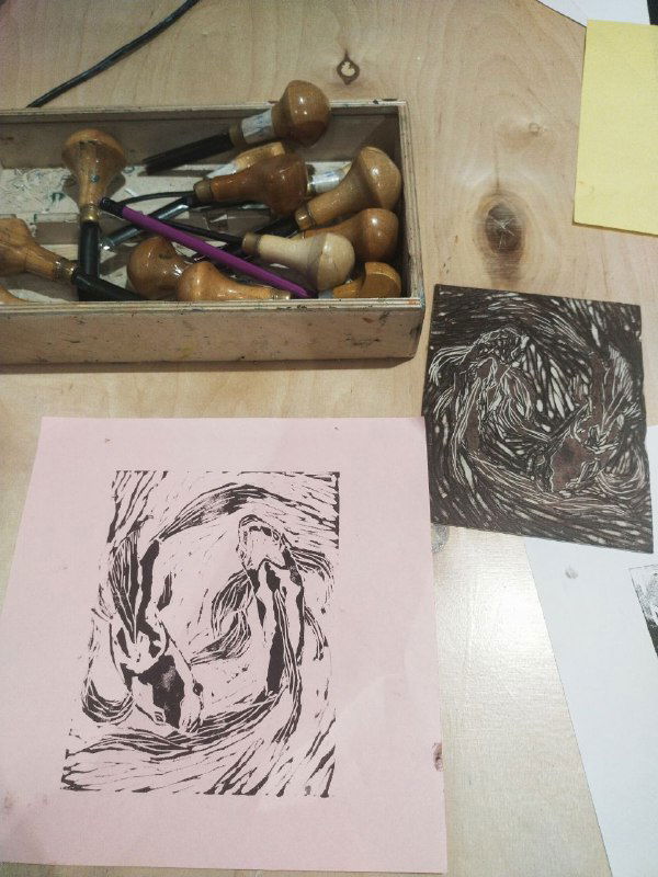 Рабочая композиция: светлый бумажный оттиск рядом с вырезанной тёмной пластиной и набором резцов и инструментов для линогравюры.