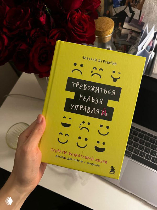 Рука держит жёлтую книгу «Тревожиться нельзя управлять» на фоне ноутбука и букета роз — фото читательницы, иллюстрация к тексту о дистанцировании эмоций.