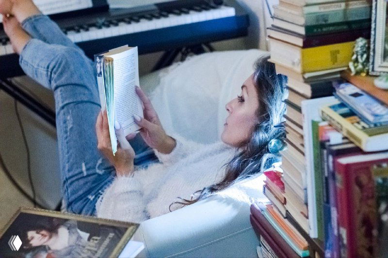 Женщина читает книгу, устроившись в кресле среди стопок книг и музыкальной клавиатуры в уютной домашней обстановке, символ заботы о себе.