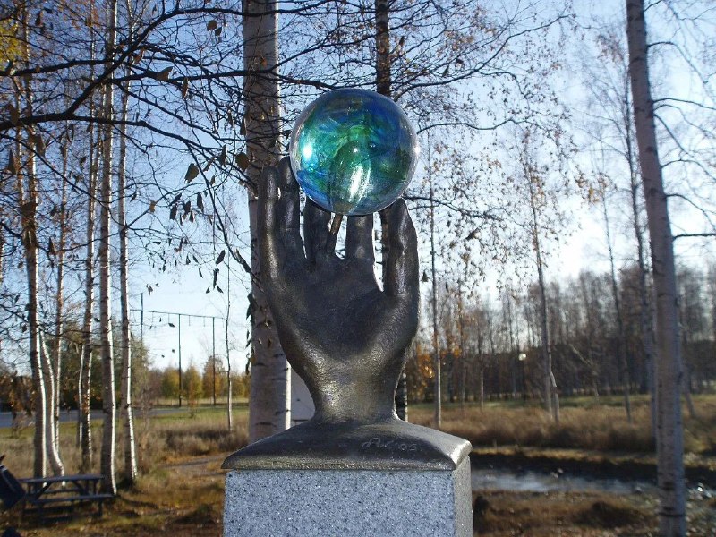 Скульптура руки, поднимающей прозрачный стеклянный шар, установлена на постаменте на фоне парковой растительности и дорожек.
