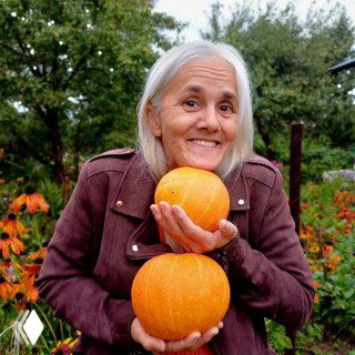 Пожилая женщина улыбается и держит две тыквы на фоне сада; фото иллюстрирует историю вдовы о жизни после утраты.
