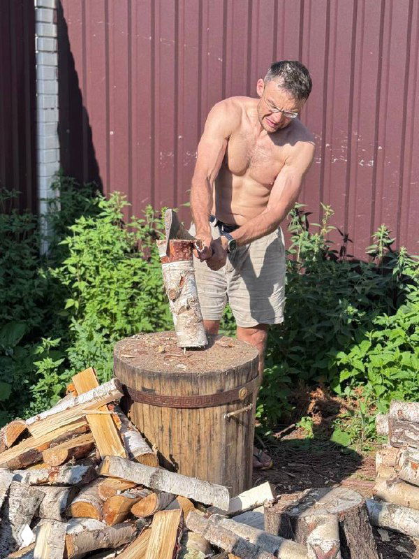 Взрослый мужчина без рубашки колет дрова на большом пне во дворе у металлического забора; рядом стопка поленьев, солнечный летний день, бытовая сцена.