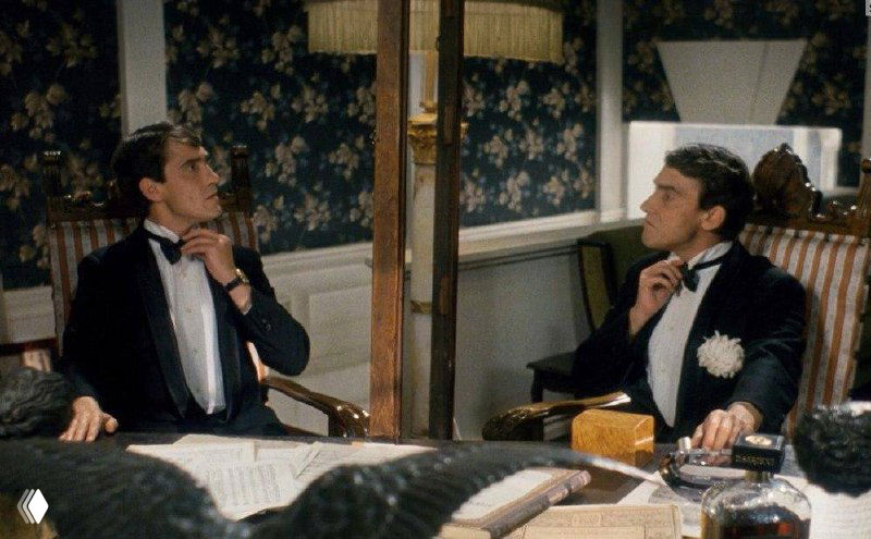 Двое мужчин в смокингах сидят за столом в комнате с узорчатыми обоями; один поправляет бабочку — иллюстративное фото для деловой истории о переговорах и статусе.