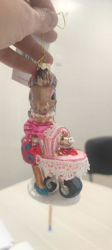 Украшение-лошадка на шнурке в руке автора, вид сбоку с деталями розовой раскраски, миниатюрной каретой и блёстками на поверхности игрушки.
