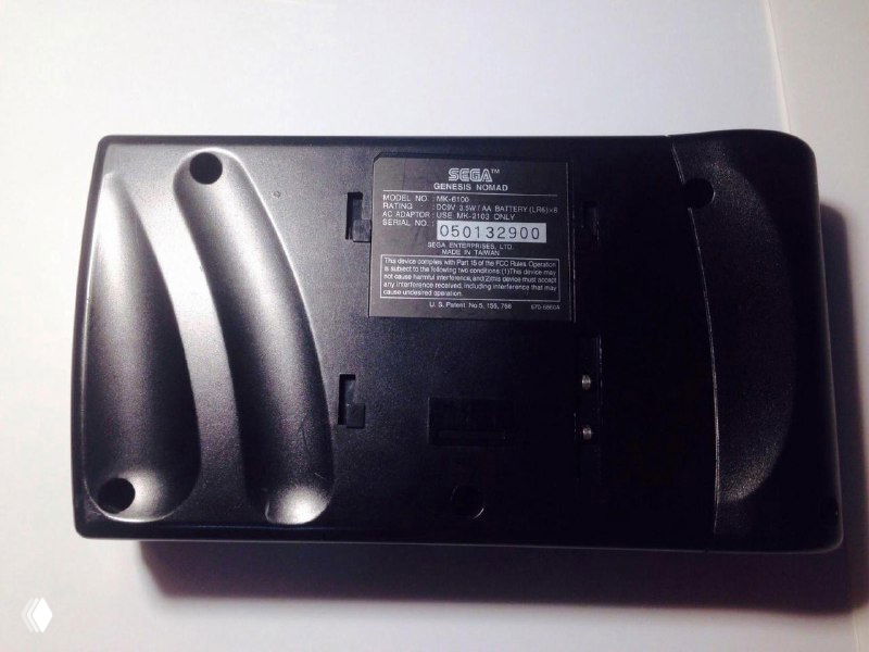 Вид снизу и сзади SEGA Nomad: пластиковый корпус, винтовые крепления и информационная наклейка с серийным номером, фото на белом фоне.