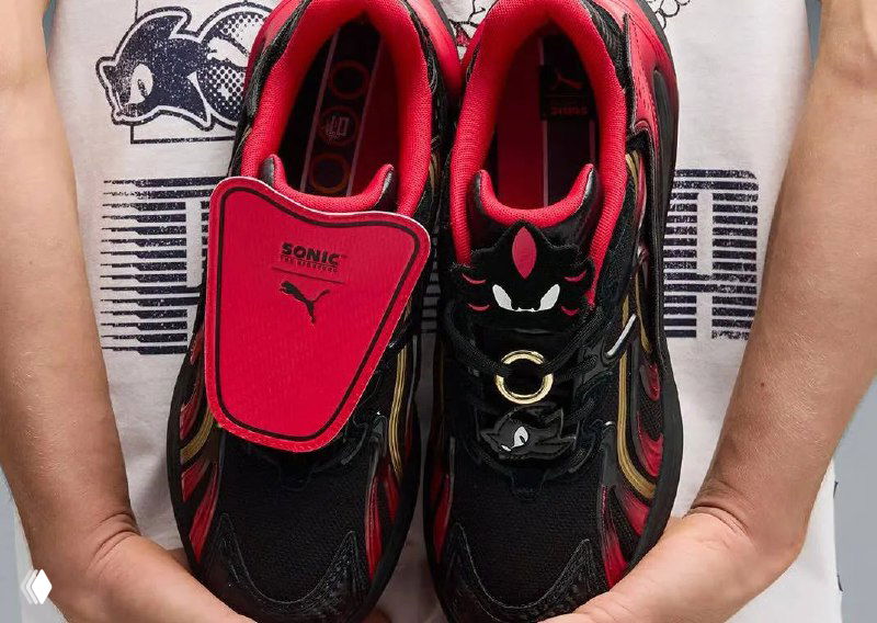 Крупный план красно-чёрных кроссовок Puma с декоративной язычковой накладкой и небольшим брендингом Sonic, вид сверху на фоне футболки с принтом.