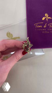 Крупный план кулона маски FUOCO из Fiore di Firenze в руках, видно оформление коробки бренда и красно‑золотые эмалевые детали изделия.