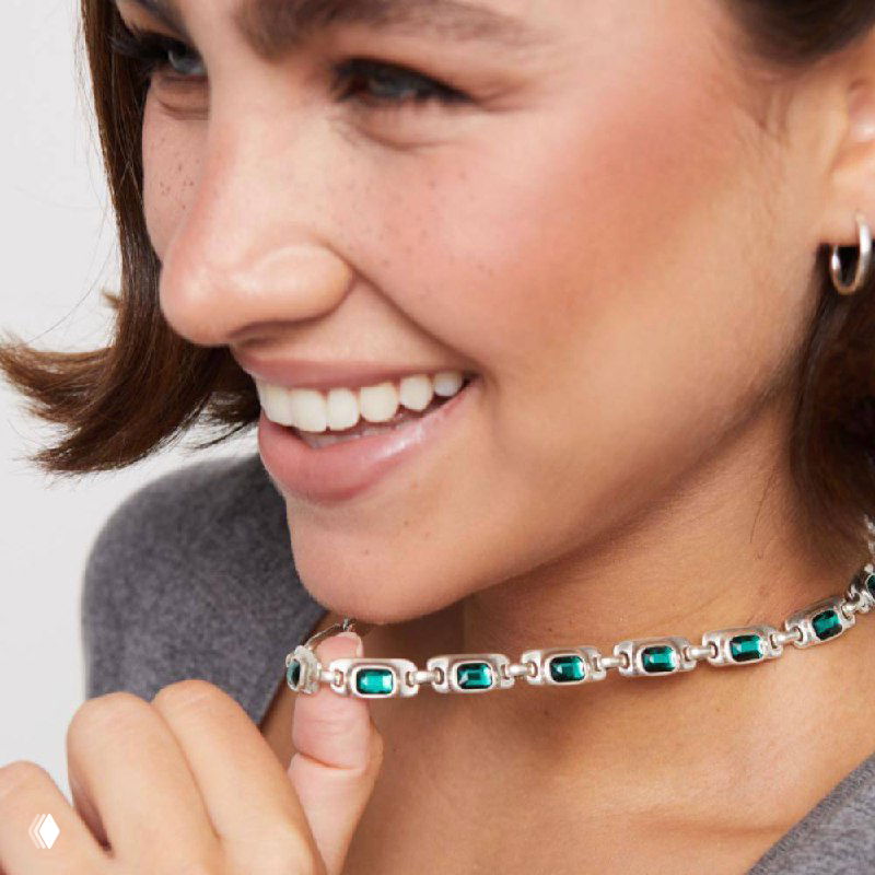 Крупный план: модель улыбается, держит колье с зелёными прямоугольными кристаллами из коллекции Ser Magnetica бренда UNOde50.