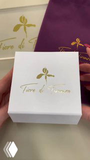 Белая подарочная коробка с золотым логотипом Fiore di Firenze на фоне фиолетового мешочка — упаковка сета украшений для презентации в магазине.