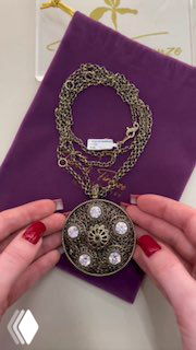 Крупный план декоративной подвески на цепях, размещённой на фиолетовом мешочке; подвеска с филигранной вставкой и круглыми розовыми элементами.