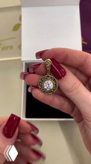 Рука с маникюром держит круглую акцентную подвеску в белой коробке — крупный план подвески с розовой центральной вставкой и декоративной окантовкой.