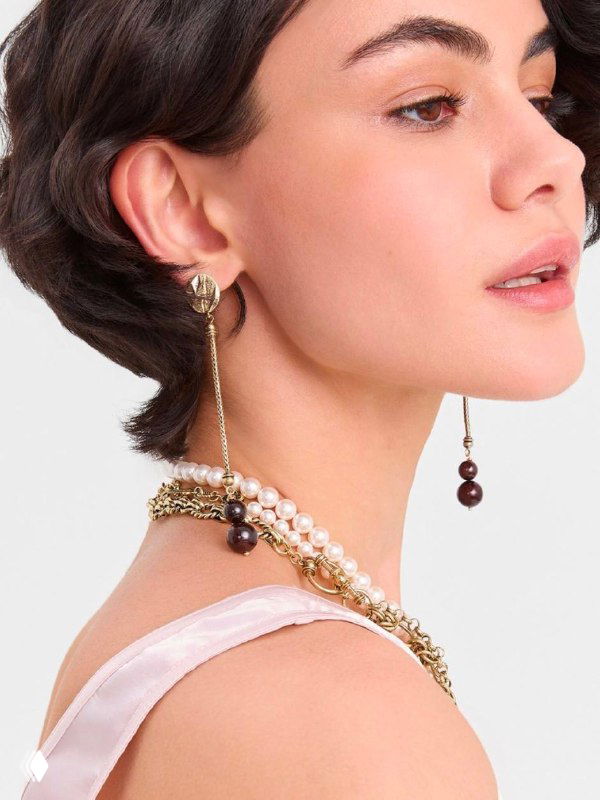 Крупный план модели с короткой стрижкой: длинные подвесные серьги, многослойные ожерелья и деликатный макияж на светлом фоне, рекламный кадр каталога.