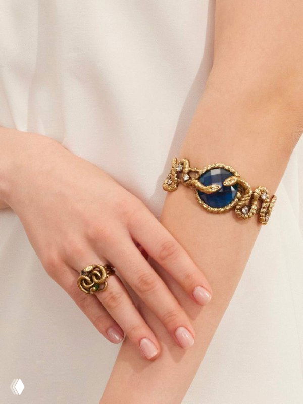 Крупный план рук и запястья с массивным браслетом и кольцом: золотистые детали и синие вставки, фокус на текстуре украшений и отделке.
