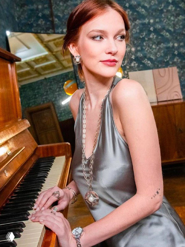 Модель в серебристом платье у рояля носит длинные цепи и серьги, кадр в ретро‑атмосфере подчеркивает элегантность украшений и вечеринную стилистику.