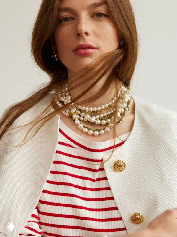 Модель в светлой одежде демонстрирует многослойное жемчужное колье и брошь: спокойный дневной образ с акцентом на классические украшения.