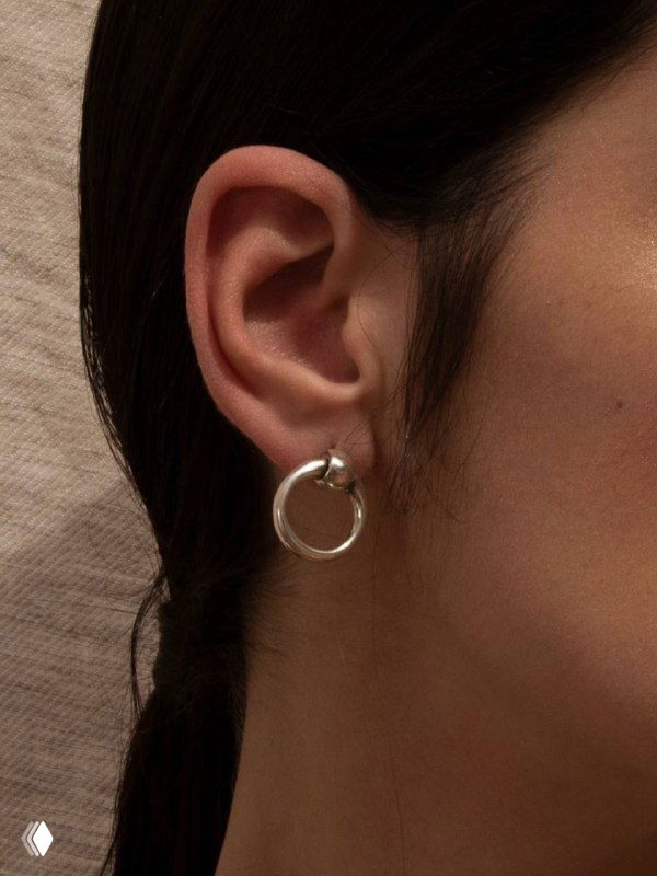 Крупный план уха в естественном освещении с небольшой серебристой серьгой-кольцом CICLON; акцент на форме и качестве покрытия металла.
