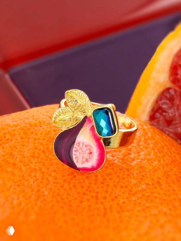 Крупный план кольца TARATATA с эмалевым изображением фрукта и разноцветным декоративным камнем на оранжевом фоне в студийной композиции.