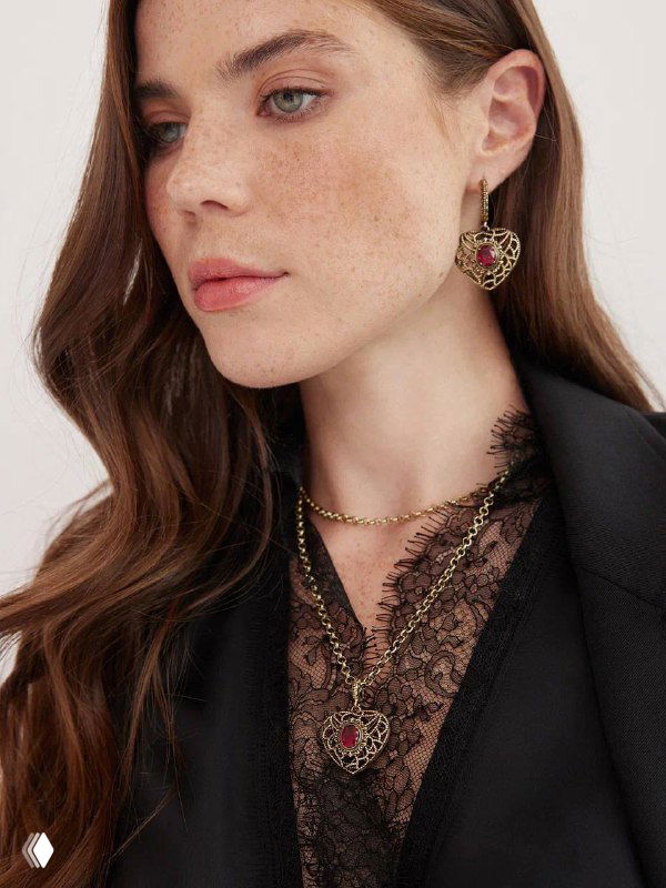 Женская модель в чёрном жакете демонстрирует золотистые серьги и кулон в форме сердца с тёмно‑красной вставкой, крупный план.