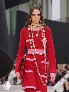 Подиумный кадр: модель в красном платье с многослойными жемчужными ожерельями, демонстрация тренда многослойности в украшениях.