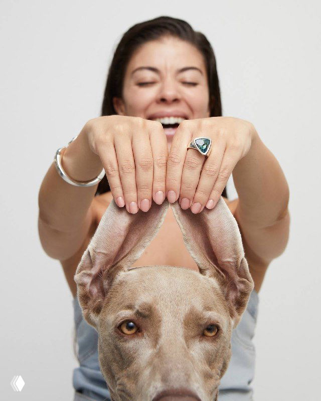 Женщина держит собаку и демонстрирует кольцо Ser Camaleonica (UNOde50) с зелёным камнем; тёплый эмоциональный фэшн-кадр с питомцем.