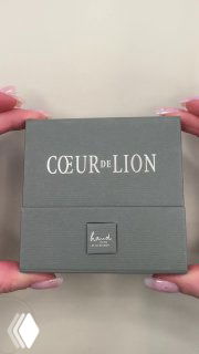 Двойной кадр серой подарочной коробки COEUR de LION в руках продавца — вид спереди с логотипом и текстурой упаковки.
