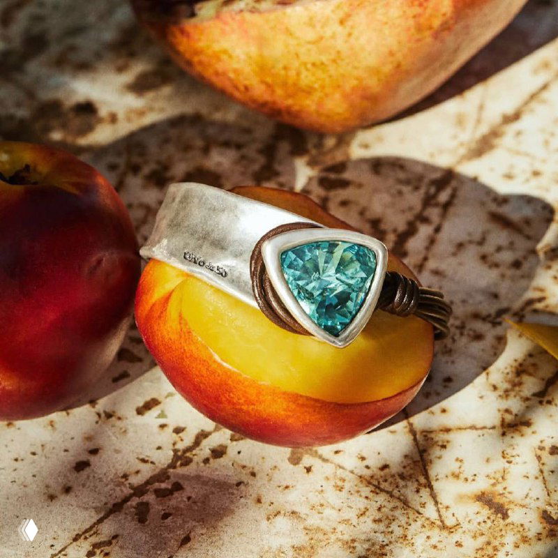 Крупный план серебристого украшения UNOde50 на фоне разрезанного фрукта; зелёный камень, текстура фрукта и тёплая подсветка в рекламной стилистике магазина.