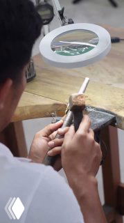 Ювелир за рабочим столом обрабатывает деталь украшения под лупой: деревянная подставка, инструменты и руки мастера в процессе ручной сборки.