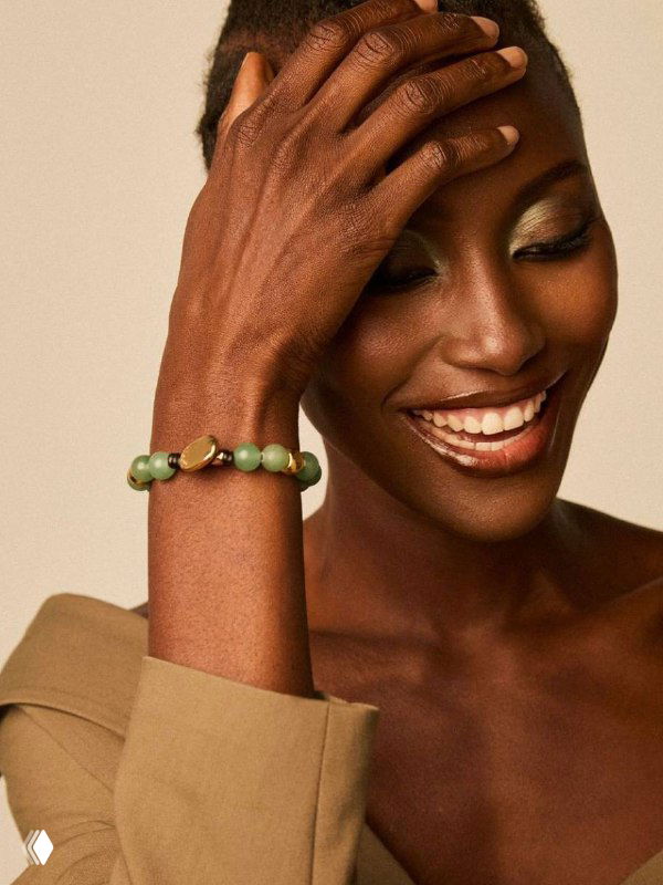 Улыбающаяся модель в бежевом образе показывает браслет VIDDA с зелёными камнями — рекламная съёмка новой летней коллекции бренда