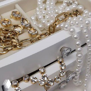 Крупный план: золотая цепь, жемчуг и кристаллы, выложенные на белой подставке — акцент на текстуре и блеске украшений.