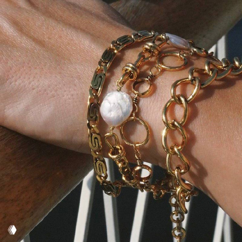 Снимок запястья с несколькими браслетами: золотые цепи разной фактуры и один жемчужный акцент — стильный многослойный комплект.