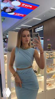 Полный рост в зеркало: женщина в голубом комплекте украшений стоит перед витриной бутика с подсветкой и дисплеем ювелирных товаров.