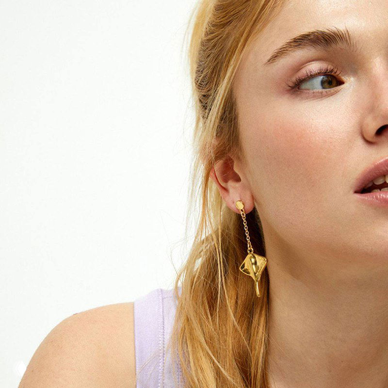 Крупный план золотистых и серебристых серёжек с мотивом морского ската на ухе модели — аксессуары из коллекции Ser Majestuosa для летних образов.