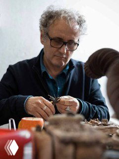 Итальянский мастер за рабочим столом держит в руках украшение, инструменты и детали, кадр передаёт процесс ручной работы и атмосферу мастерской