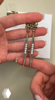 Крупный план длинных серьг в форме меча из коллекции IL SPADA, держатся на пальцах — видны белые кристаллы и металлическая текстура.