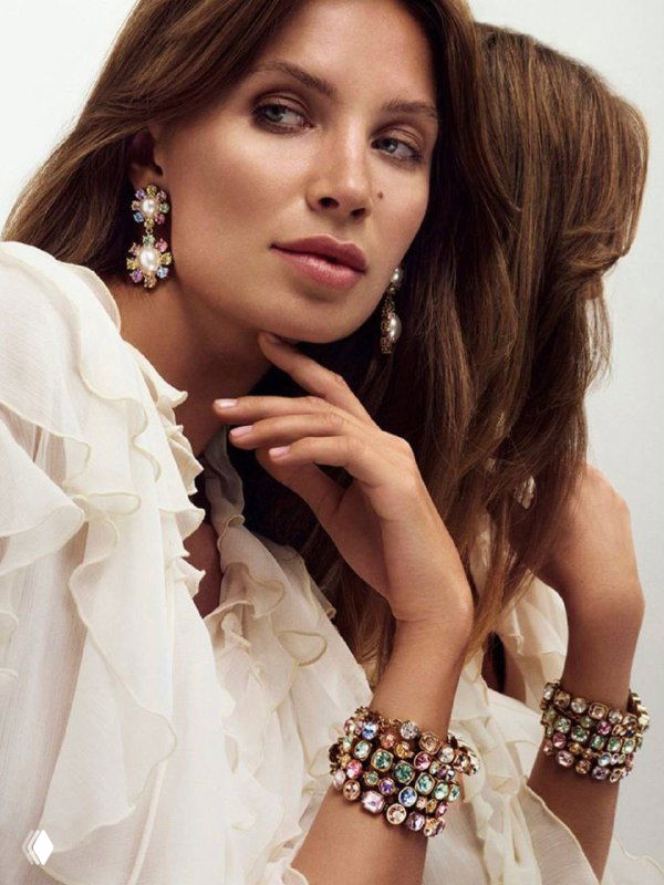 Женская модель в белом платье демонстрирует серьги и браслеты Dyrberg Kern с крупными разноцветными камнями в студийной рекламной съёмке.