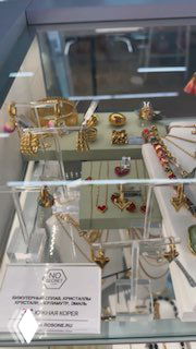 Витрина магазина ROSONE: золотистые и позолоченные украшения бренда NO SECRET расставлены на подставках в торговом зале ТЦ Discovery, видна часть стеклянной витрины и отражение пола.