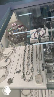 Дисплей с серебристыми цепочками, кольцами и серьгами бренда NO SECRET: видны разноцветные вставки, стильные колье и серьги в витрине магазина ROSONE рядом с аксессуарами.