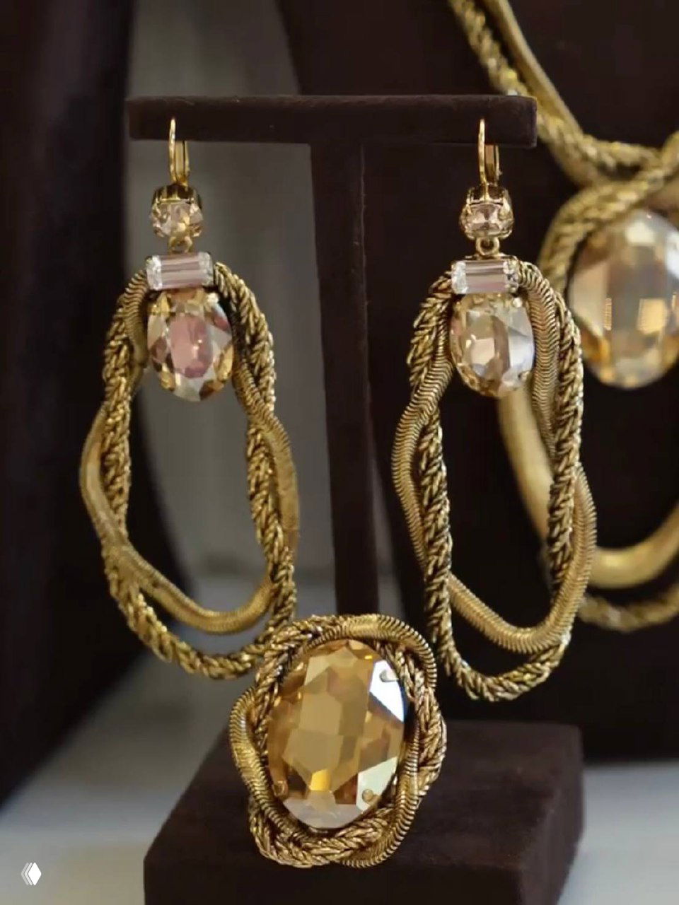 Крупный план крупных овальных и прямоугольных кристаллов в оправе и плетёных золотистых элементах — акцентный комплект Claudio Canzian на подставке.