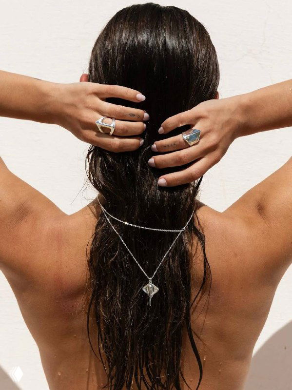 Вид сзади: украшения UNOde50 на шее и пальцах модели, длинные волосы и многослойные цепочки подчёркивают стиль коллекции.