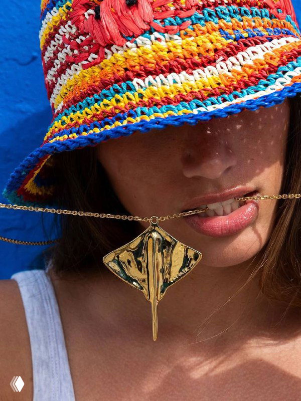 Портрет в яркой шляпе: модель держит золотистую подвеску-скат из коллекции SER MAJESTUOSA у лица на фоне насыщенных цветов.
