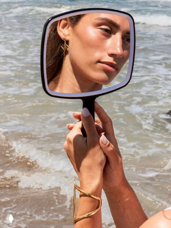 Модель у моря смотрит в ручное зеркало; серьги и браслет из коллекции SER MAJESTUOSA дополняют пляжный образ и подчёркивают летнюю тематику.