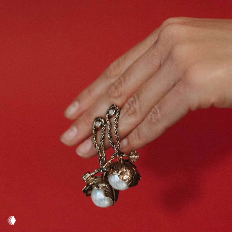 Рука держит серьги с крупными жемчужинами и декоративными подвесками на красном фоне, демонстрация фактуры, цепочек и композиций декора.